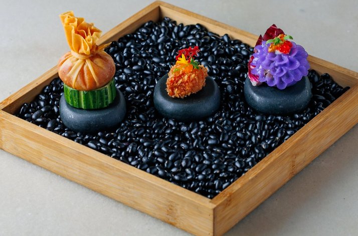 Three ornate Thai canapes displayed on black stones nestled in dark beans within a bamboo wooden tray.