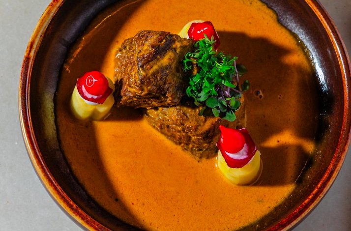Braised meat resting in rich orange curry sauce, garnished with fresh microgreens and red-capped potato bites.