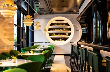 Le Jardinier Miami dining room with emerald green chairs, a lit circular herb wall feature, and a dark bar area.