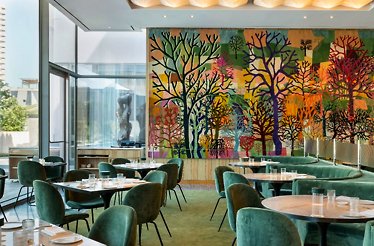 Sunlit dining room with green velvet seating and a colorful tree mural at Le Jardinier Houston.