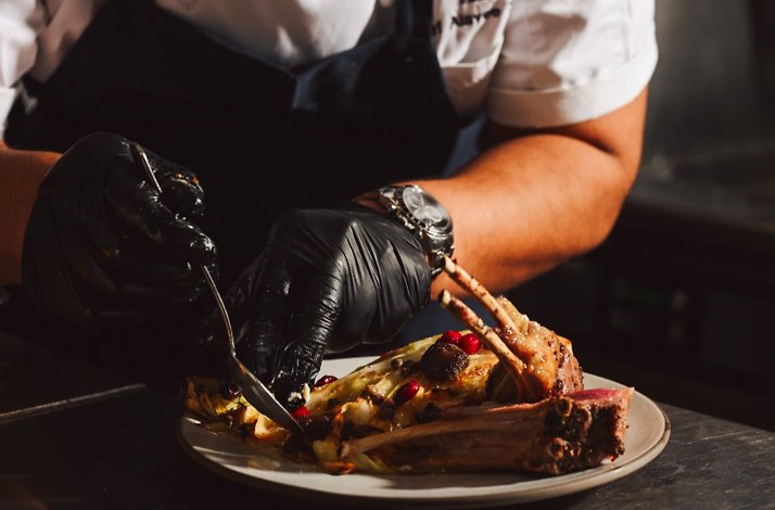 A chef wearing black gloves cutting a roasted rack of lamb served on a white plate.