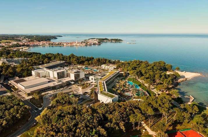 Aerial view of a seaside resort complex surrounded by pine trees, pools, and a calm bay with a coastal town beyond.
