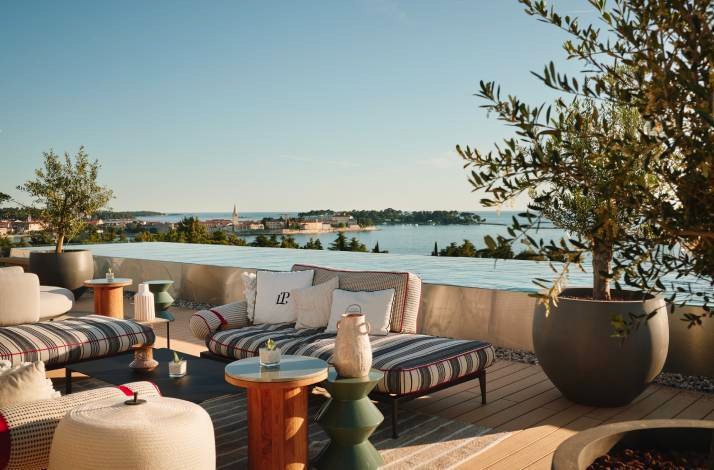 Rooftop lounge with striped daybeds, small tables, and potted trees overlooking the sea and a distant coastline at sunset.