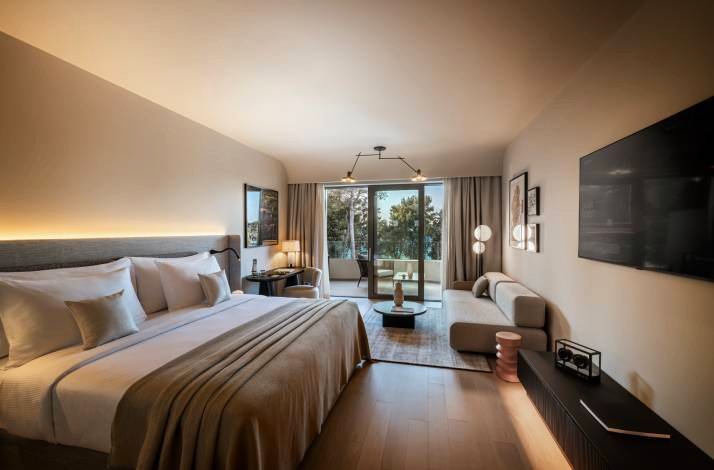 Modern hotel suite with a king bed, sofa, and balcony doors opening to treetop and sea views in soft evening light.