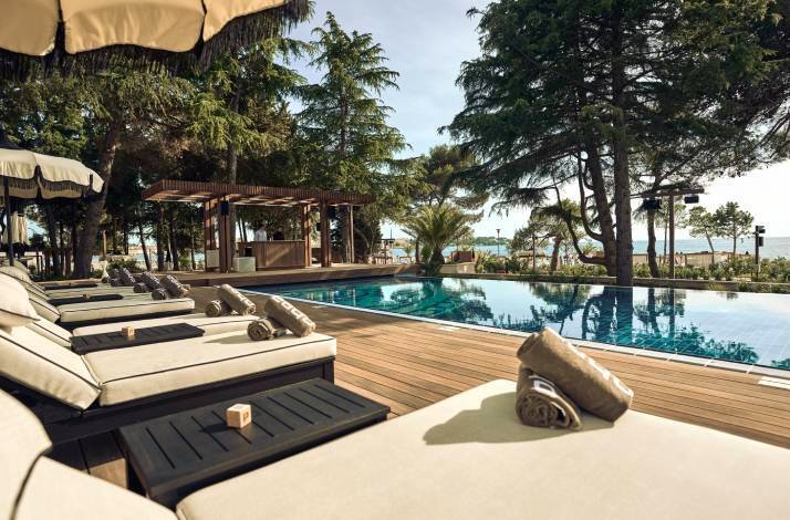 Poolside sun loungers with rolled towels beside an infinity-style pool shaded by trees, with the sea visible in the distance.