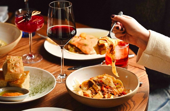 A hand lifting wide-ribbon pasta from a bowl surrounded by red wine, cocktails, focaccia, and shared plates at Porto Boston.