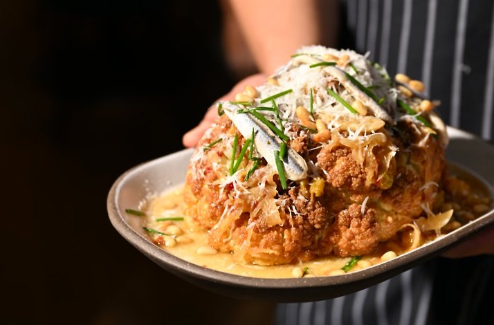 Whole roasted cauliflower topped with shaved cheese, pine nuts, anchovies, and chives, held on a plate by a chef at Porto Boston.