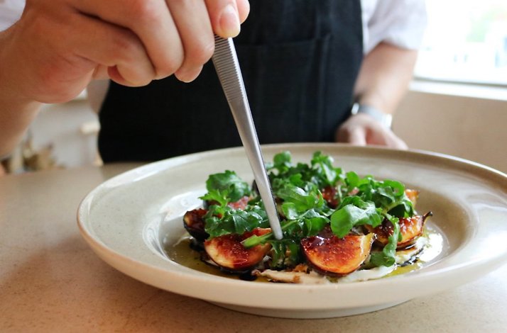 Chef using tweezers to plate roasted figs with fresh greens and cream on a ceramic dish at Osteria Giulia.