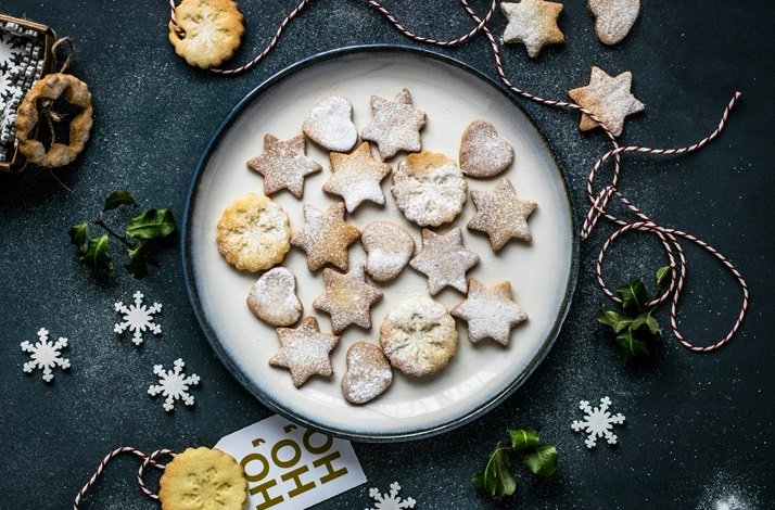 Star, heart, and round shortbread cookies dusted with powdered sugar on a white plate, with holly and snowflakes nearby.