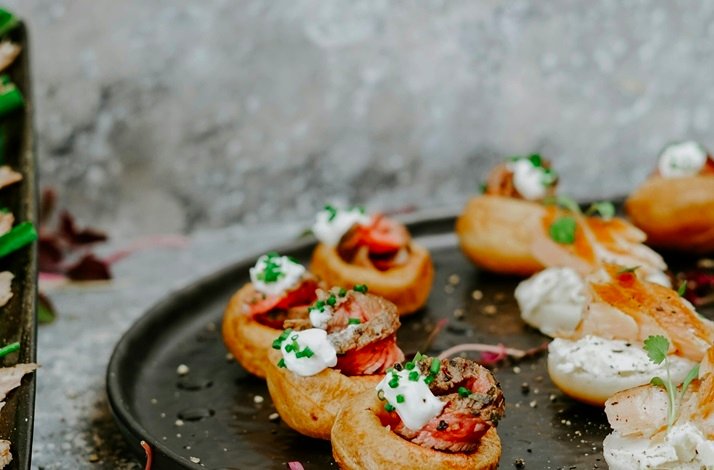 Bite-sized Christmas canapés with sliced pink meat, cream, and chives on crispy pastry cups on a dark plate.