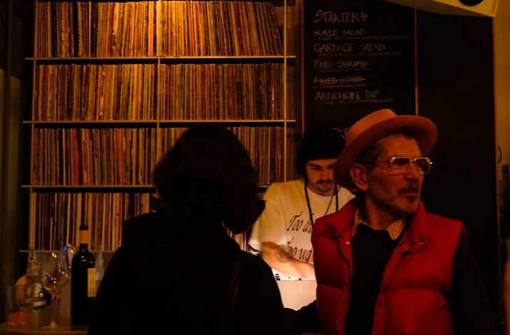 Dim bar with vinyl wall; people near counter, man in hat and red vest looks to the side.