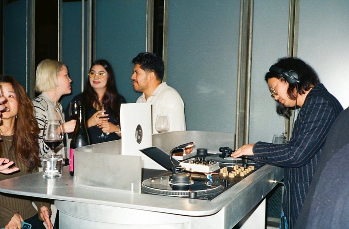 DJ mixes at a turntable while guests chat and drink wine at a small bar counter.