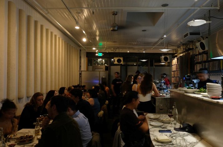 Busy restaurant dining room with long marble bar, seated guests, and warm overhead lighting.