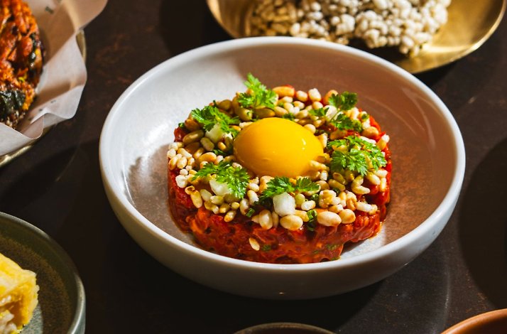 Korean beef tartare topped with a golden egg yolk, pine nuts, and fresh herbs, served in a shallow white bowl.