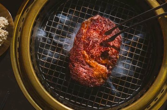 Sizzling dry-aged beef cut held by tongs on a round charcoal grill, with smoke curling upward at Takja. 