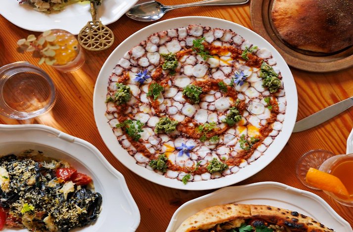 Overhead spread of dishes including sliced octopus carpaccio with harissa and borage flowers, black pasta and flatbread.