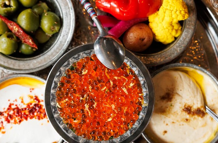 Overhead view of silver mezze bowls filled with salmon roe, green olives, hummus, and pickled vegetables.