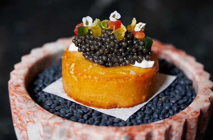 Golden fried potato cake topped with black caviar, cream, diced peppers, and edible flowers, served in a pink marble vessel.