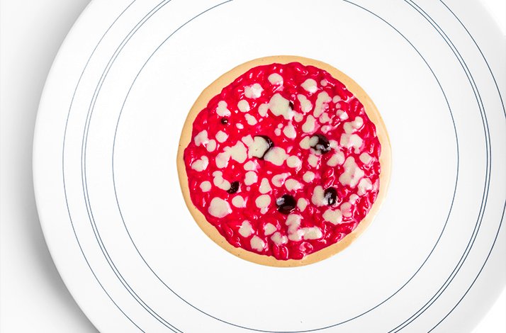A white plate holds a circular dish of vibrant pink risotto drizzled with white sauce and scattered dark berries.
