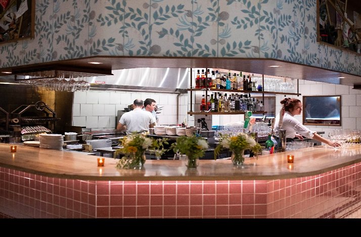 Open kitchen bar with pink tile counter, bottles on shelves, and staff working at L’Oca d’Oro.
