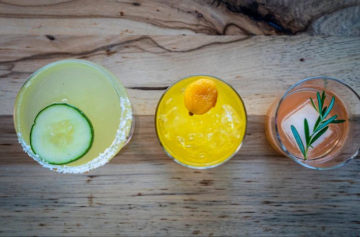 Trio of garnished cocktails on a wood surface, featuring cucumber, kumquat, and rosemary herb sprigs as finishing touches.