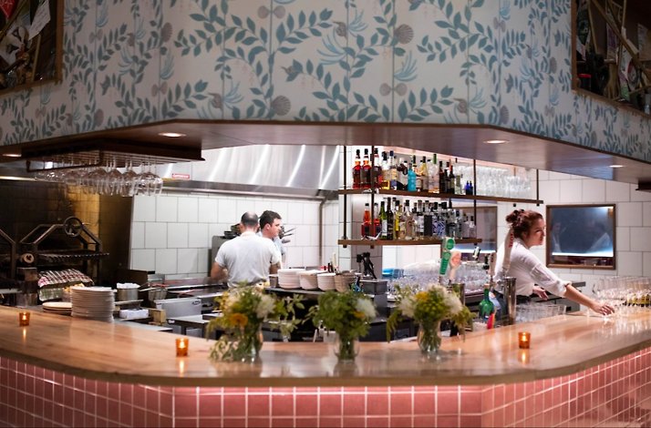 Open restaurant kitchen and bar with staff working, liquor shelves, hanging glassware, and fresh florals on a wooden counter.