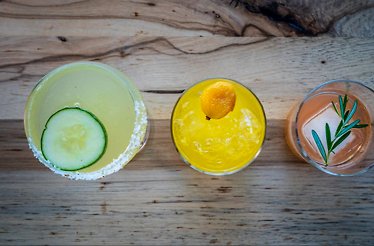 Trio of garnished cocktails on a wood surface, featuring cucumber, kumquat, and rosemary herb sprigs as finishing touches.