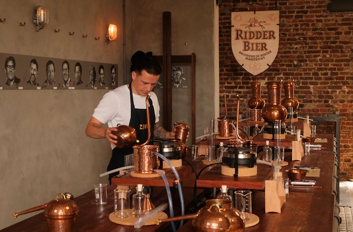 ZIVIO instructor operating copper gin stills at City Distillery Maastricht, with a 'Ridder Bier' heritage logo displayed on an exposed brick wall.