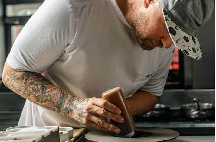 Chef Jeremy Ford focused on plating a dish in a restaurant kitchen while wearing a white apron and cap.