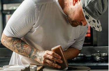 Chef Jeremy Ford focused on plating a dish in a restaurant kitchen while wearing a white apron and cap.
