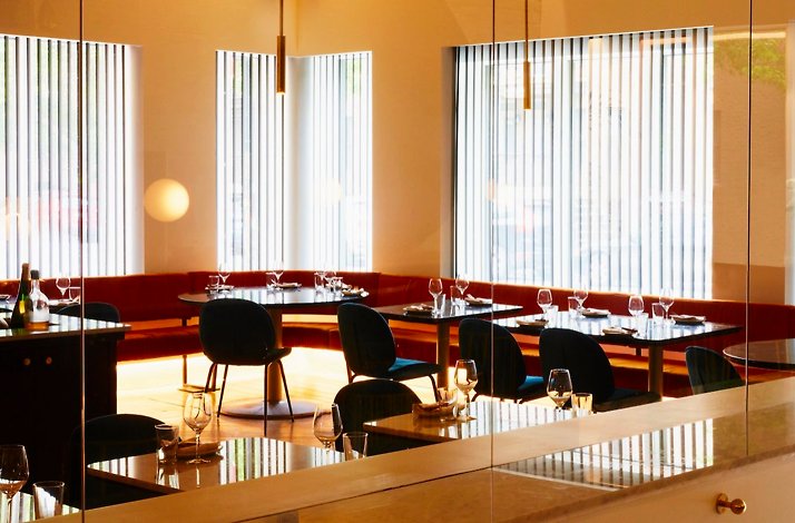A warm ambience dining interior of Elena Restaurant, showcasing red banquette seating, black tables, and tall sunlit windows. 
