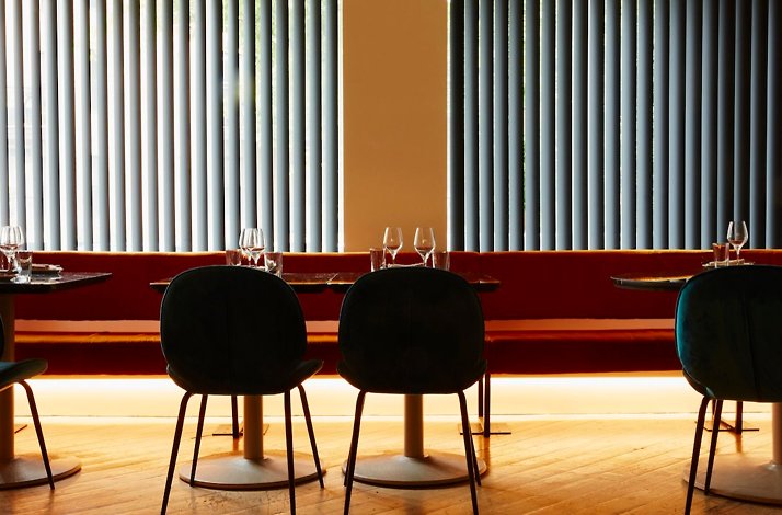 A warm ambience dining interior of Elena Restaurant, showcasing red banquette seating, black tables, and tall sunlit windows. 