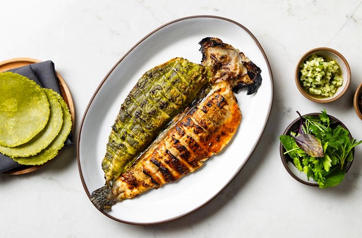 Image 2: A large platter of dual-marinated grilled fish served with green tortillas, chopped salsa, and fresh herbs.