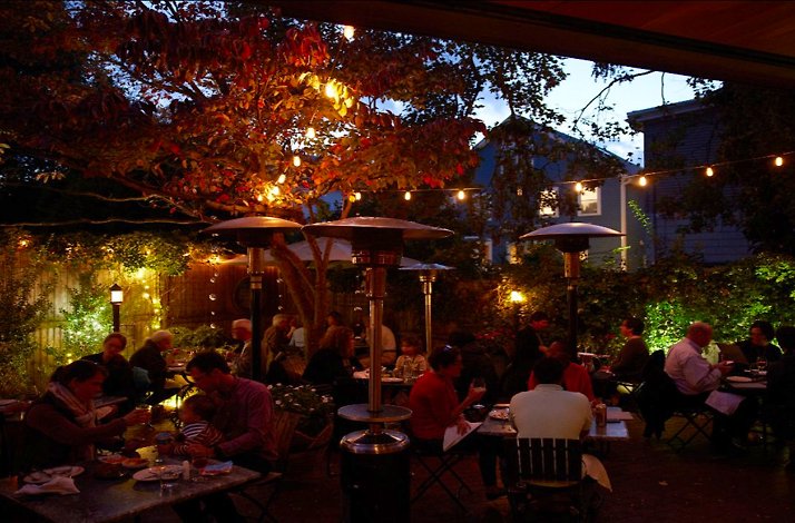 Oleana Restaurant's outdoor patio at dusk filled with dining guests beneath warm string lights, glowing trees, and patio heaters. 