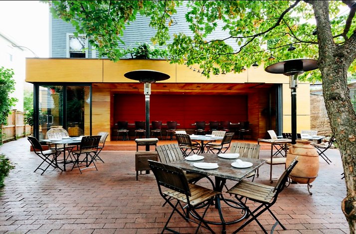 Oleana Restaurant's brick patio featuring set dining tables, bistro chairs, patio heaters, and a warm wood-paneled exterior.