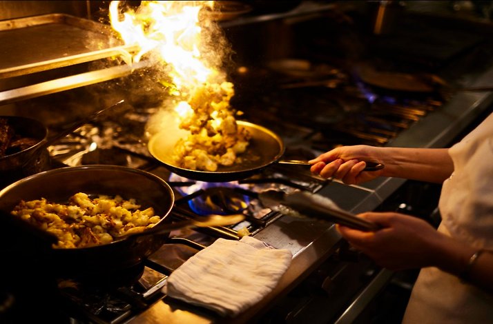 Chef's hands flambeing cauliflower in a skillet over a gas flame in Oleana Restaurant's kitchen, with a second pan nearby.
