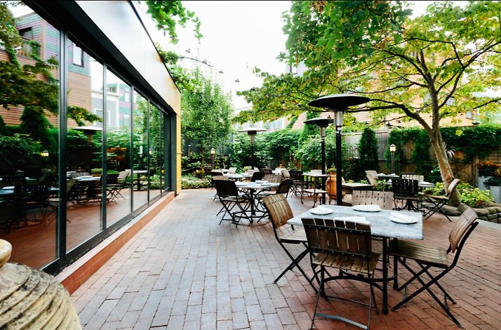 Oleana Restaurant's garden patio with set tables, bistro chairs, patio heaters, and lush surrounding greenery beside glass-walled facade.