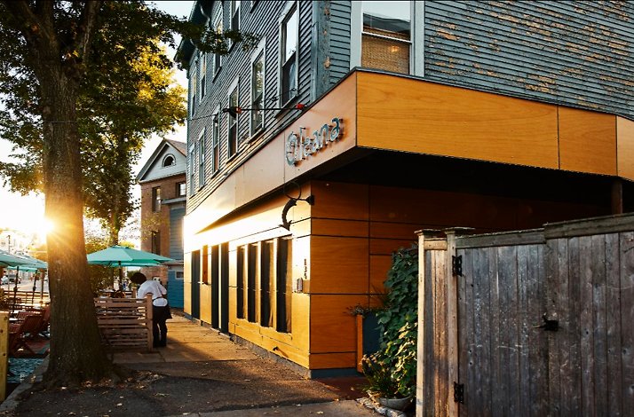 Oleana Restaurant's wood-paneled exterior at sunset, featuring the 'Oleana' sign and a server heading toward the sidewalk patio.