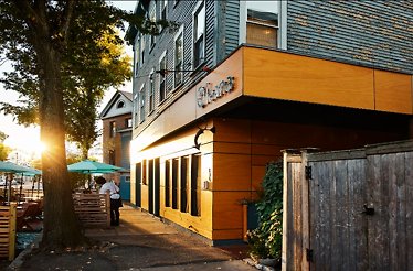 Oleana Restaurant's wood-paneled exterior at sunset, featuring the 'Oleana' sign and a server heading toward the sidewalk patio.