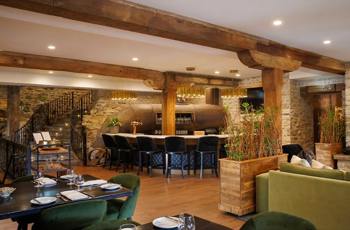 Elora Mill Hotel dining room featuring exposed wood beams, stone walls, a curved bar, green velvet seating, and set tables.