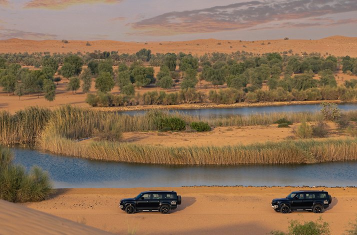 Two black Land Rovers traversing the expansive desert landscape of the Dubai Desert Conservation Reserve, with a river in view.